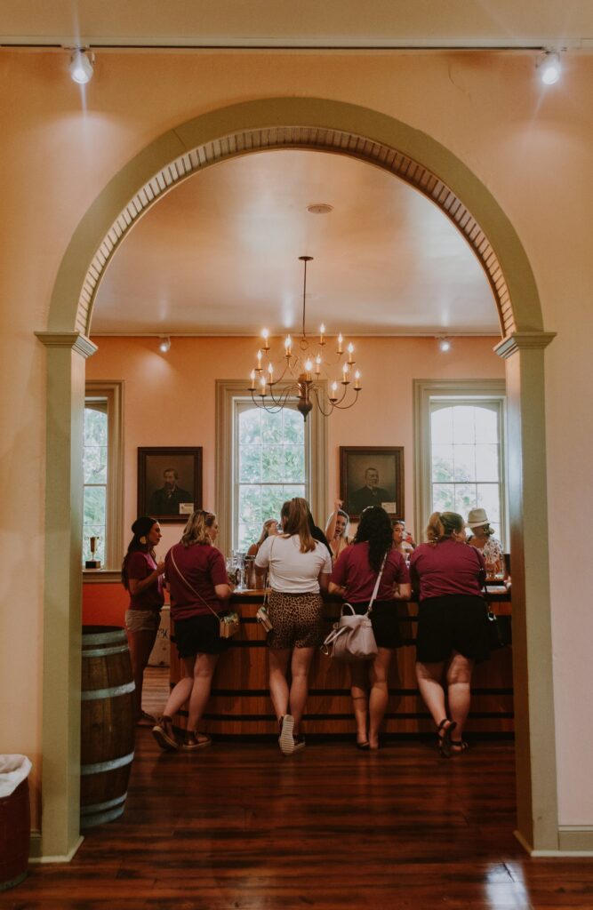 Guests tasting wine at the bar in the Founder's Tasting Room, seen through an arched doorway under a chandelier
