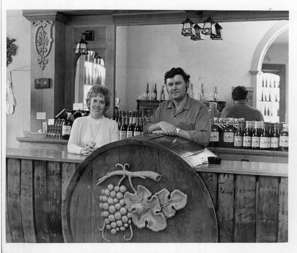 Jim and Betty Held behind the bar in the tasting room, with a carved grape cluster barrelhead in front